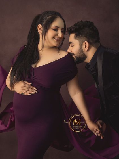 A playful and loving moment between a couple. The partner gently kisses his wife's shoulder as she smiles, dressed in a beautiful purple gown with flowing fabric.