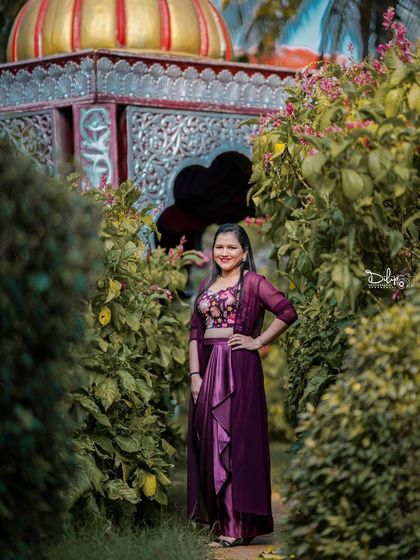 A solo portrait of a model in a beautiful purple dress, posing in a garden pathway with a gazebo in the background.