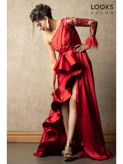 A model looking stunning in a red gown with an intricate one-shoulder design. Her hair is styled in a chic, braided updo, perfect for a formal event or red carpet.