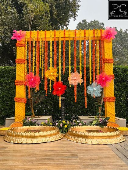 This outdoor Haldi setup features a bright yellow draped frame with hanging marigold strings and colorful paper flowers. The two large brass urlis are ready for the bride and groom's ceremony.