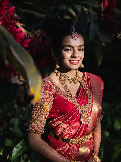A smiling bride in a classic red bridal saree. The blouse is heavily embroidered with gold thread, creating a look of timeless elegance.