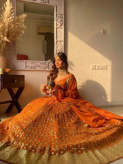 A beautiful seated pose in the orange lehenga, perfect for a festive photoshoot. The rich color and gold details create a warm and inviting look.