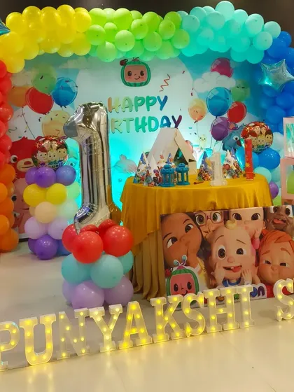 A vibrant and colorful Cocomelon theme for a first birthday party. This setup features a rainbow balloon arch, character cutouts, and large marquee letters spelling out the birthday girl's name.