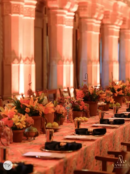 A long, communal dining table set for a feast, with a runner of terracotta pots filled with orange flowers, set against the grand arches of the palace.