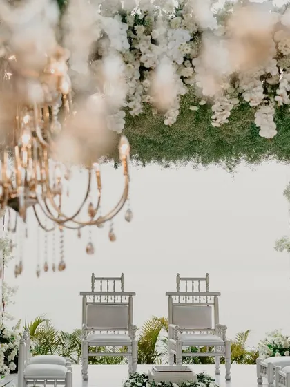 A detailed shot of the mandap seating, framed by a cascade of white flowers and a crystal chandelier. This view highlights the intricate floral work and the serene, dreamlike quality of the all-white theme.