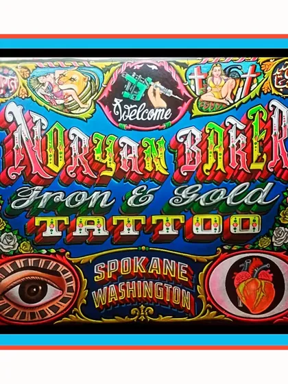A sign for Noryan Baker's Iron & Gold Tattoo in Spokane, Washington. This piece is full of tattoo-related imagery like an eye, a heart, a tattoo machine, and religious symbols, all framed in an ornate border.