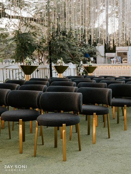 Modern and sophisticated seating for an engagement party. The dark, textured chairs with gold legs are arranged neatly, facing a bar area adorned with cascading string lights.