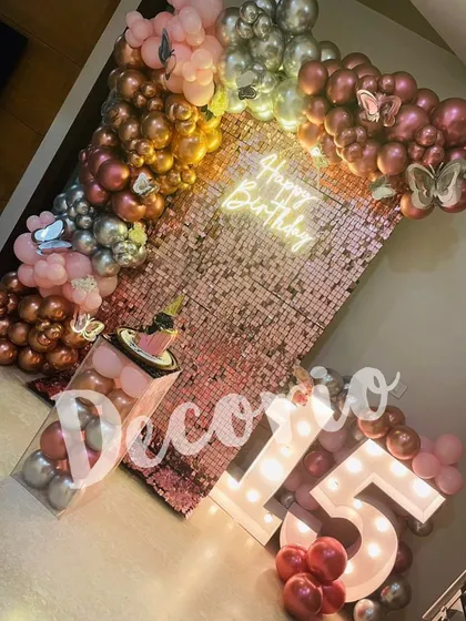 A stylish 15th birthday setup featuring a rose gold sequin wall. The decoration is enhanced with marquee numbers, a neon sign, and a pink and metallic balloon arrangement.