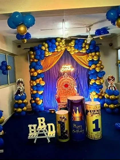 A grand and royal prince-themed first birthday party. The decor is rich with royal blue and gold balloons, a curtained backdrop, a throne for the birthday boy, and custom-themed props.