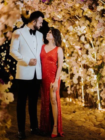 The couple shares a look of love under a tunnel of warm, glowing flowers. The groom's white tuxedo and the bride's sparkling red dress create a look of pure Hollywood glamour.