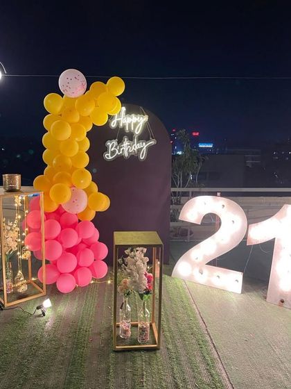 This is a duplicate of ID 21, 60, 91, 108, 121, 168, 206, 232, 239. A vibrant 21st birthday decoration on a rooftop. This setup features a balloon arrangement in hot pink and yellow, a 'Happy Birthday' neon sign, and large marquee numbers against a city skyline.