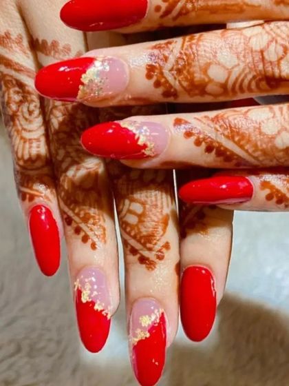 A bold and beautiful design for a festive occasion, pairing vibrant red nails with a sheer base and gold leaf accents, complementing the client's henna.