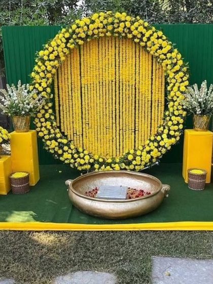 A classic Haldi backdrop for outdoor functions. This setup features a large circular frame filled with strings of yellow marigolds, a green panel background, and a traditional brass urli in the center for the ceremony.
