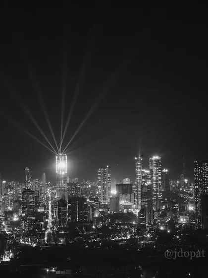 A black and white photograph of the Mumbai skyline at night during Diwali, with spotlights from the Palais Royale creating a dramatic effect.