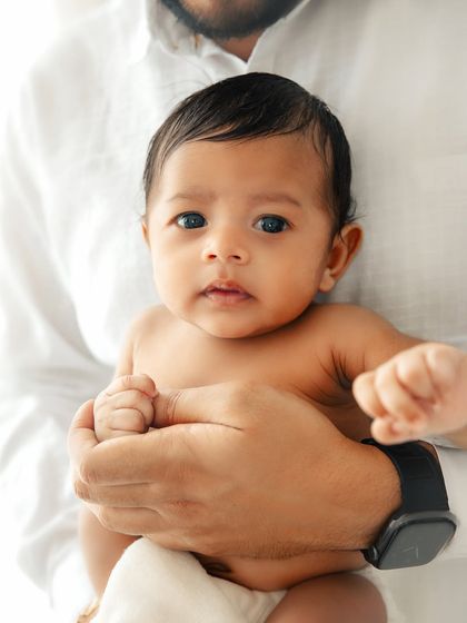 A close-up portrait of a newborn's face, held securely in his father's hands. The focus is on his beautiful eyes and delicate features.