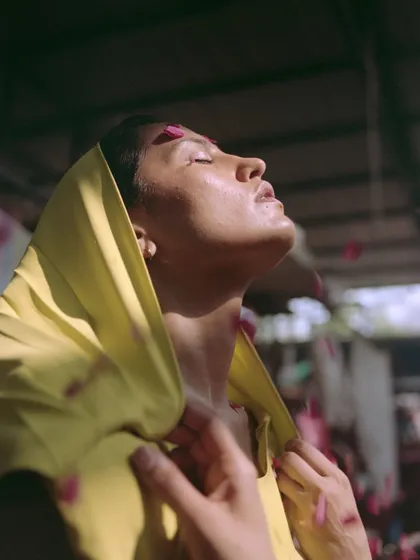 An ethereal portrait where the model is bathed in sunlight, wearing a hooded yellow dress. The styling creates a serene, almost spiritual moment amidst the chaos of the flower market, highlighted by a shower of pink petals.
