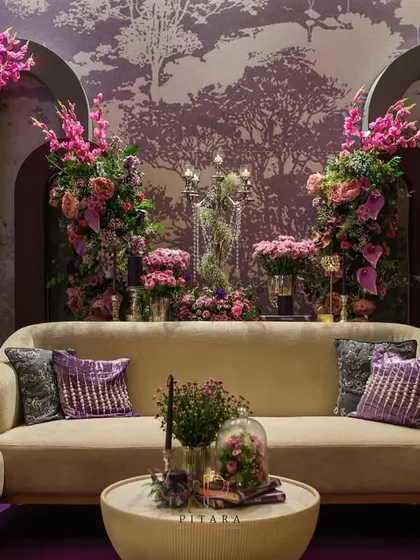 The central seating area of the lounge, featuring a classic cream sofa with plush purple cushions. The floral arrangements framing the sofa add a sense of depth and luxury.