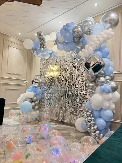 A dreamy birthday setup with a silver shimmer wall, a 'Happy Birthday' neon sign, and a beautiful balloon arch in blue, white, and silver, designed to look like clouds.