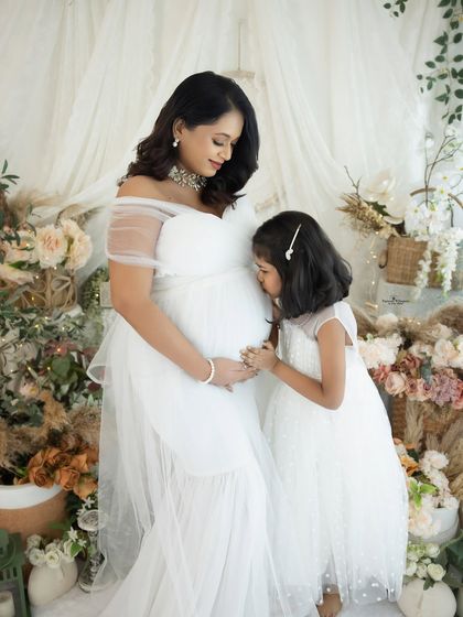 A precious moment between a mother and daughter, both in white. The daughter gently kisses her mother's baby bump, a beautiful symbol of sibling love.