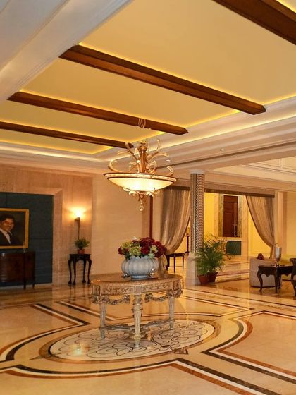 In this grand hall, wooden ceiling beams and Art Nouveau flooring create a harmonious backdrop. The interplay of beige marble, dark wood, and a statement chandelier establishes a sense of traditional luxury.
