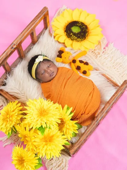 A newborn wrapped in an orange swaddle sleeps in a miniature wooden bed, surrounded by bright yellow sunflowers. This setup is perfect for a warm, sunshine-filled portrait.