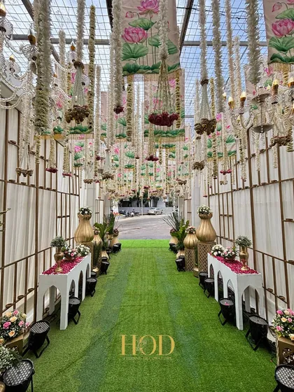 A long walkway with a green turf carpet, lined with traditional decor elements and leading to a grand entrance under a ceiling of hanging flowers and chandeliers.