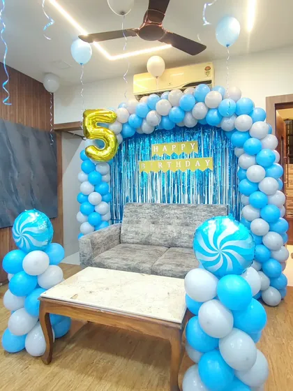 A classic blue and white balloon arch for a 5th birthday. This home setup includes a shimmer curtain backdrop, a 'Happy Birthday' banner, and two candy-swirl foil balloons on pillars.