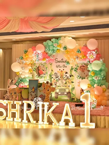 A vibrant jungle-themed first birthday stage for Shrika. The setup includes marquee letters, a lush balloon arch in pastel colors, and adorable cutouts of jungle animals like a giraffe and lion.