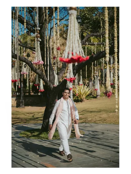 The groom's stylish solo walk at the Haldi venue, with beautiful floral decorations hanging from the trees, setting a festive and elegant scene.