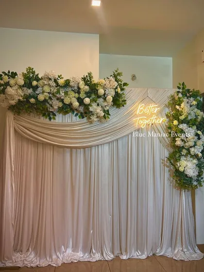 This elegant Roka backdrop uses asymmetrical floral arrangements on a white draped wall. The 'Better Together' neon sign adds a personal touch, making it perfect for capturing memories of your special day.
