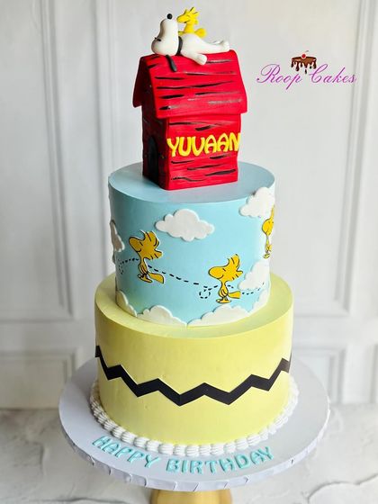 A charming three-tier Snoopy themed cake. The design features the iconic zig-zag pattern, Woodstock flying in the clouds, and is topped with a fully edible fondant doghouse with Snoopy on top.