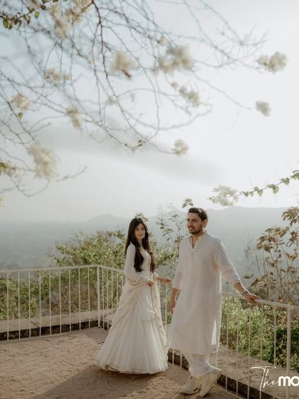 A pre-wedding portrait with a stunning natural backdrop. The couple stands on a balcony overlooking a valley, their matching white outfits creating a beautiful contrast with the lush landscape.