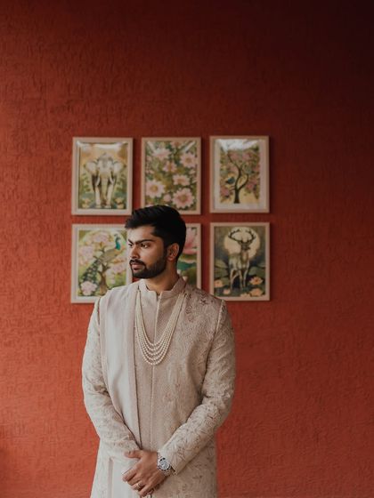 A stylish portrait of the groom against a warm, terracotta-colored wall with art frames. His elegant sherwani and confident pose create a modern yet traditional look.