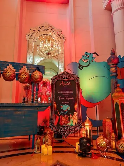 A whimsical welcome setup featuring a large Genie cutout, a themed cart, and a collection of golden lanterns and props.