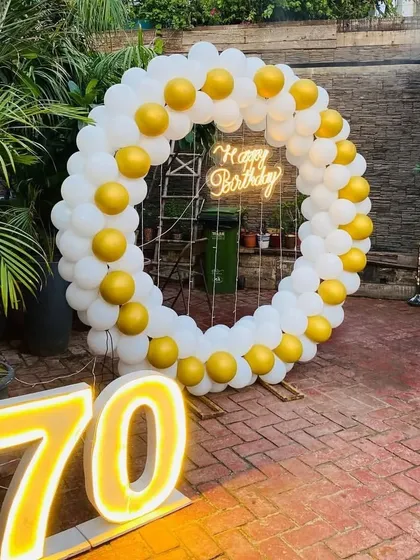 A beautiful outdoor setup for a 70th birthday, with a white and gold balloon ring, a neon sign, and large '70' marquee numbers.