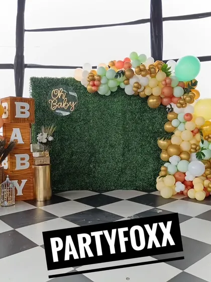 A rustic-chic baby shower decoration with a green hedge wall, wooden "BABY" blocks, and an arch of muted green, orange, and gold balloons.