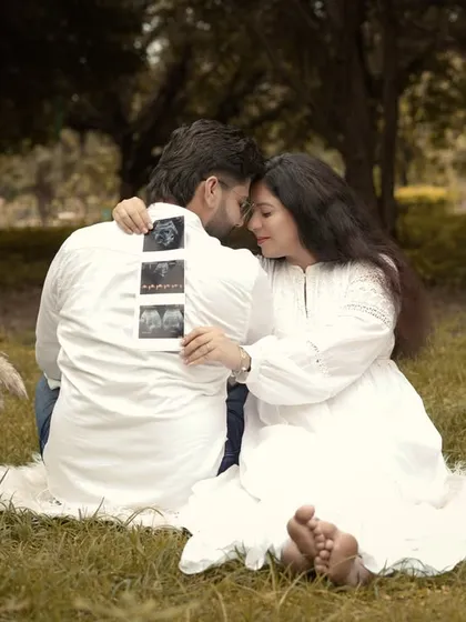 A sweet and intimate moment during an outdoor picnic-style shoot. The couple, dressed in white, shares a quiet moment while holding up their baby's first pictures, the sonogram.