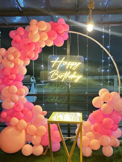 A vibrant pink balloon semi-arch on a rooftop. The neon 'Happy Birthday' sign glows beautifully against the evening sky, creating a perfect party atmosphere.