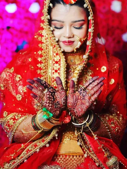 A stunning bride showing her hands in prayer. The dark mehendi stain stands out beautifully against her red bridal outfit, completing her look perfectly.