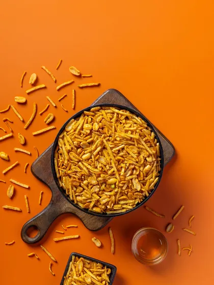 A spicy mixture photographed from above for Sartaj Sweets & Namkeen. The bright orange background complements the warm tones of the snack, creating an energetic product shot.