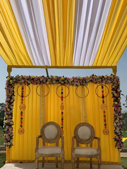 A close-up of the outdoor Haldi stage. The backdrop is decorated with floral hoops and hanging elements, framed by a border of colorful flowers.