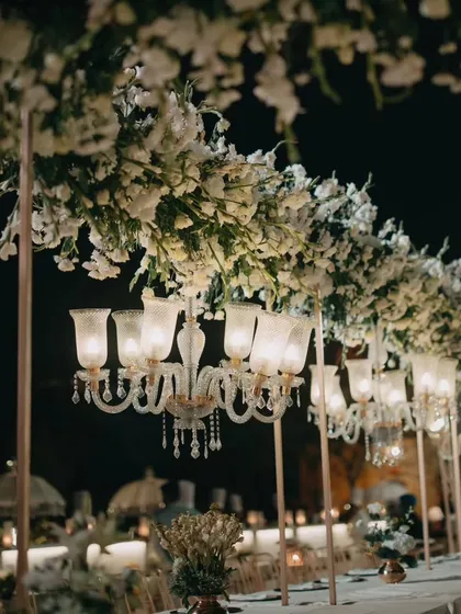 An elegant nighttime dining setup with long tables, overhead floral arrangements, and classic glass chandeliers. The ambiance is one of timeless sophistication and romance.