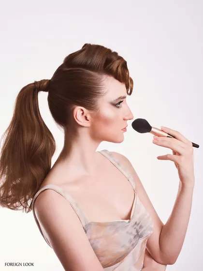 A profile view from a beauty editorial, showcasing the intricate retro hairstyle and the application of makeup with a brush.