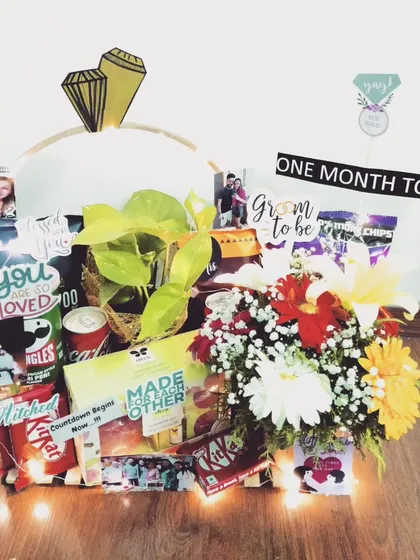 An engagement countdown hamper to celebrate the journey to 'I do'. This gift includes snacks, a live plant, fresh flowers, and is decorated with photos and fairy lights for a magical touch.