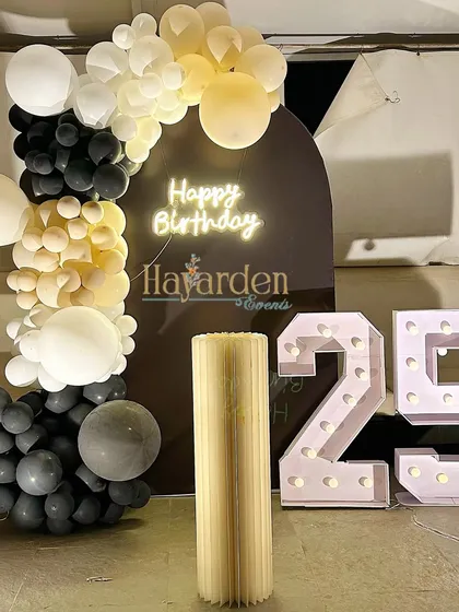 A sophisticated 25th birthday setup with a dark arched backdrop, a 'Happy Birthday' neon sign, and large marquee numbers. The black, white, and muted yellow balloon garland adds a modern, elegant touch.