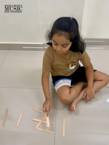 Another young learner concentrates on arranging popsicle sticks to represent a rhythm pattern. We encourage creative, tactile methods to reinforce musical learning.