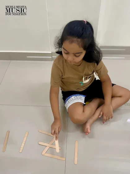 Another young learner concentrates on arranging popsicle sticks to represent a rhythm pattern. We encourage creative, tactile methods to reinforce musical learning.