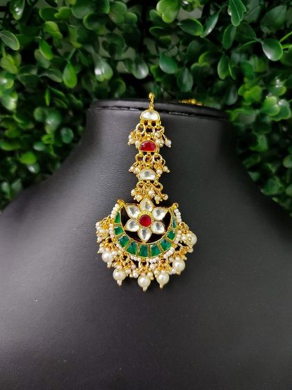 A lovely crescent-shaped maang tikka with a floral kundan motif and red and green stones.