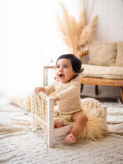 A happy baby girl sitting on a miniature bed in a simple, light-filled boho setup. Her joyful expression is everything.