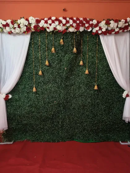 For clients wanting a simple yet elegant traditional backdrop, I offer designs like this one. It combines a lush green wall with classic white drapes, a floral header of red and white roses, and hanging temple bells for a touch of grace suitable for any small ceremony.